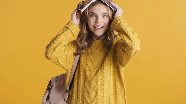陽気なブロンドティーンの学生の女の子開催本上の頭の上に本を見て喜び黄色の背景に孤立しました おかしい生徒の女の子 — ストック写真
