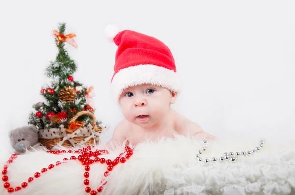 Niedliches Baby mit Weihnachtsmütze und einem Weihnachtsbaum dahinter — Stockfoto