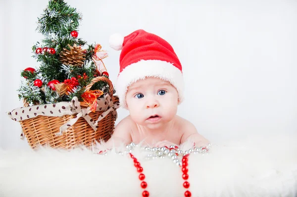 Niedliches Baby mit Weihnachtsmütze und einem Weihnachtsbaum dahinter — Stockfoto