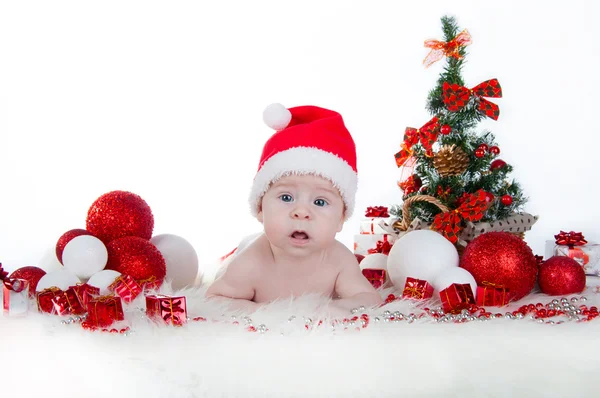 Niedliches Baby mit Weihnachtsmütze und einem Weihnachtsbaum dahinter — Stockfoto