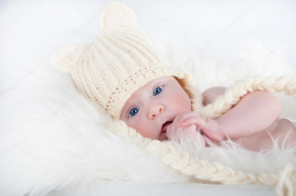 A cute little baby is looking into the camera and is wearing a white hat. The baby could be a boy or girl and has blue eyes.