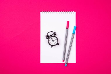 Kayıtlar için beyaz bir defter, iki farklı renk sapı kırmızı zemin üzerinde küçük bir siyah alarm saati. Ofis içeriği.