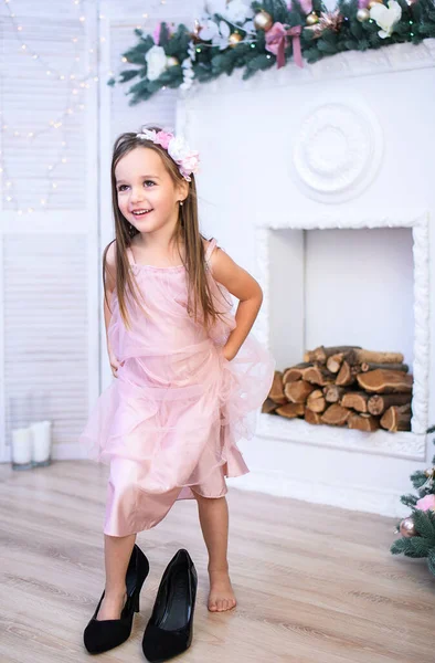 우아 한 벽난로 근처의 아름다운 분홍 드레스를 입은 한 어린 소녀가 어머니의 굽 굽을 바라봅니다. 새해 카드. — 스톡 사진