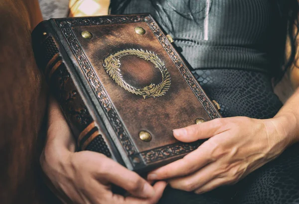 Siyah elbiseli bir kız kanepeye oturuyor ve elinde yaldızlı sembollü kahverengi deri bir kitap, kabartmalı çerçeve ve kitap omurgasında kaburga kaldırıyor..