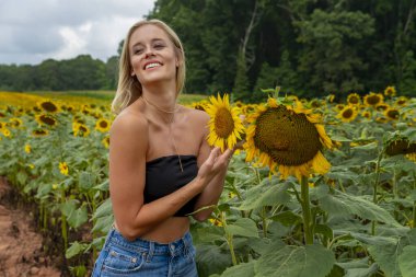 Sarışın bir model, yaz gününün tadını çıkarırken ayçiçeği tarlasında poz veriyor.