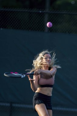 Sarışın bir model tenis kortunda öğleden sonra güneşinde tenis oynuyor.