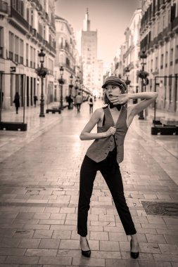 Çekici Avrupalı bir kadın hareketli bir İspanyol caddesinde kendine güvenen bir poz vererek, kıvrımlarını şık bir kıyafetle ortaya koyuyor. Güneşli arka plan ve hareketli atmosfer onun şık ve zahmetsiz zarafetini ön plana çıkarıyor..