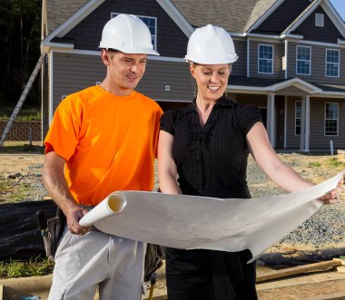 Güneşli bir inşaat sahasında, bir inşaat işçisi ve bir mühendis birlikte planları inceliyor, çizgileri ölçüyor ve ayrıntıları tartışıyor çerçeveler yükselirken, takım çalışması güvenli bir aile evini şekillendiriyor..