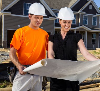 Güneşli bir inşaat sahasında, bir inşaat işçisi ve bir mühendis birlikte planları inceliyor, çizgileri ölçüyor ve ayrıntıları tartışıyor çerçeveler yükselirken, takım çalışması güvenli bir aile evini şekillendiriyor..