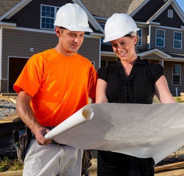 Güneşli bir inşaat sahasında, bir inşaat işçisi ve bir mühendis birlikte planları inceliyor, çizgileri ölçüyor ve ayrıntıları tartışıyor çerçeveler yükselirken, takım çalışması güvenli bir aile evini şekillendiriyor..