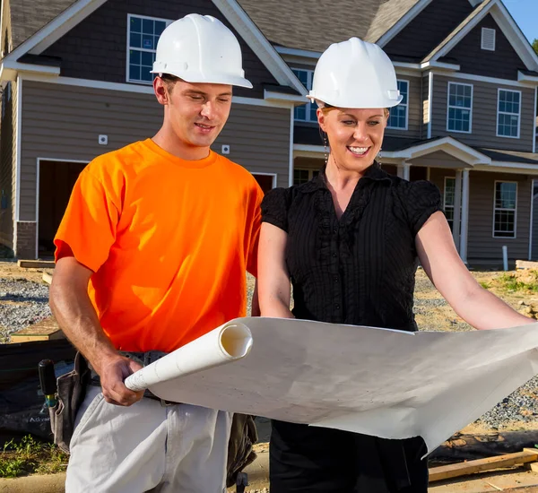 Güneşli bir inşaat sahasında, bir inşaat işçisi ve bir mühendis birlikte planları inceliyor, çizgileri ölçüyor ve ayrıntıları tartışıyor çerçeveler yükselirken, takım çalışması güvenli bir aile evini şekillendiriyor..