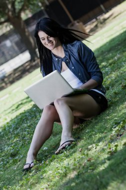 Güzel, genç bir esmer güneşli bir parkta oturuyor ve üniversite derslerine çalışırken dizüstü bilgisayarına odaklanıyor. Etrafı yeşilliklerle çevrili, öğrenmeyi doğanın huzuruyla harmanlıyor..