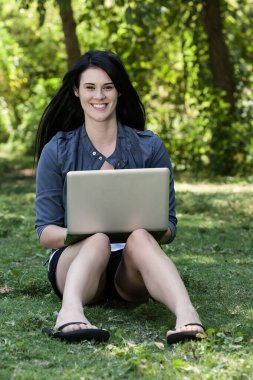 Güzel, genç bir esmer güneşli bir parkta oturuyor ve üniversite derslerine çalışırken dizüstü bilgisayarına odaklanıyor. Etrafı yeşilliklerle çevrili, öğrenmeyi doğanın huzuruyla harmanlıyor..