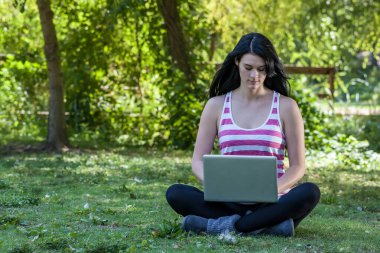 Güzel, genç bir esmer güneşli bir parkta oturuyor ve üniversite derslerine çalışırken dizüstü bilgisayarına odaklanıyor. Etrafı yeşilliklerle çevrili, öğrenmeyi doğanın huzuruyla harmanlıyor..