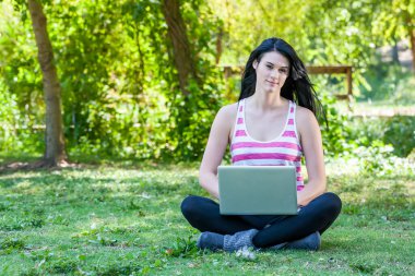 Güzel, genç bir esmer güneşli bir parkta oturuyor ve üniversite derslerine çalışırken dizüstü bilgisayarına odaklanıyor. Etrafı yeşilliklerle çevrili, öğrenmeyi doğanın huzuruyla harmanlıyor..