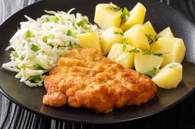 Leh usulü domuz pirzolası Kotlet Schabowy haşlanmış patates ve lahana salatasıyla birlikte siyah ahşap bir masada yakın plan tabakta. Yatay