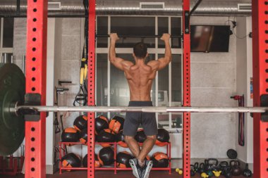 Çıplak gövdeli bir sporcunun fotoğrafı. Atletik bir adam kendini barın üzerine çekiyor. Spor salonunun çerçevesinde çeşitli toplar, halterler ve halterlerle