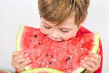 Kırmızı tişörtlü beş yaşında bir çocuk sulu bir karpuz yiyor.