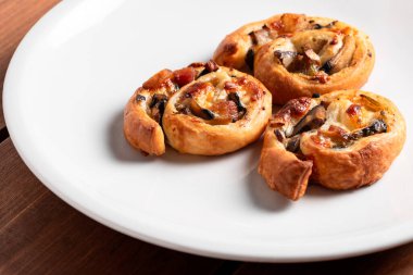 Erimiş peynir ve koyu malzemelerle dolu üç lezzetli pizza rulosu ahşap yüzeyde duran beyaz tabakta duruyor..