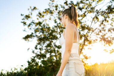 Yaz tarlasında güneş ışığı. Doğada yürüyen bir kadın sakin ve huzurlu, iyi bir ruh hali içinde. Sabah güneşi ya da akşam gün batımı. Refah ve sağlıklı yaşam tarzı tatilde. Ağaç ve mavi gökyüzü.
