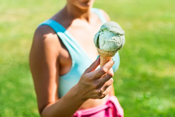Ice cream cone in hand of a fit woman in summer. Gelato and colorful fashion in park. Retro vibes in outfit and icecream on sunny weekend. Eating and enjoying summertime vacation outside. Green grass.