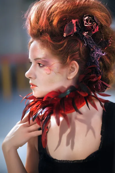 Young beautiful girl in the form of the evil fairy with red hair in a designer dress with a long skirt in unusual shoes and a necklace of feathers — Stock Photo, Image