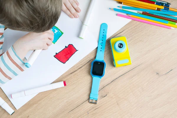 Smart baby watches with wiretapping, remote monitoring and GPS tracker lying on the table near the kid in the kindergarten. The boy draws in the album with felt-tip pens.