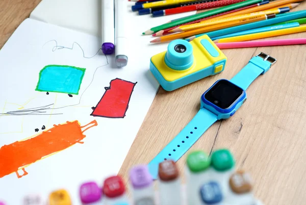 Smart baby watches with wiretapping, remote monitoring and GPS tracker lying on the table near the album for drawing, toy camera, markers and pencils. Remote control of the child concept.