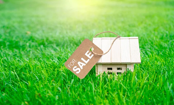 Concept of the sale of an environmentally friendly house built from natural building materials. Little wooden toy house with tag For sale on the green grass.
