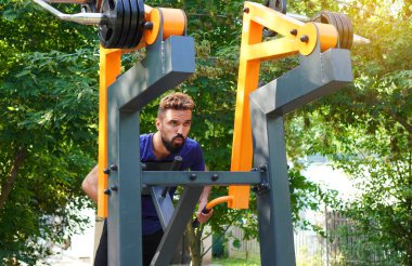 Bir adam sırtındaki kasları parktaki bir eğitim aletine pompalıyor..