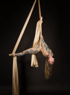 Bej takım elbiseli ince bir kız altın ipekle jimnastik ve sirk egzersizleri yapıyor. Karanlık bir arkaplanda stüdyo çekimi, izole görüntüler.