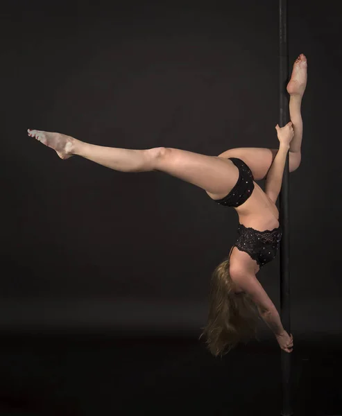 Athletic girl with long hair performs exercises on a portable pole in a black swimsuit. Studio shooting performances on a pole on a black background.