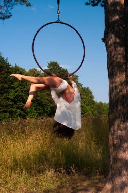Beyaz sahne kostümlü bir kız açık havada havadaki bir hava halkası içinde egzersiz yapıyor, ormanın ve gökyüzünün arka planına karşı. Sokakta fotoğrafçılık.