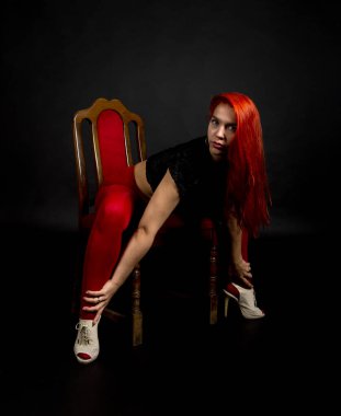 Kırmızı taytlı genç bir kadın ve uzun saçlı siyah bir yelek siyah bir arka plana karşı sandalyede poz veriyor. Stüdyo çekimi.