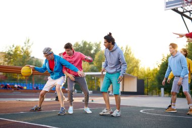 Gençler dışarıda basketbol oynuyor, oyun parkında eğleniyorlar.