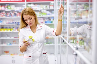 Üniformalı sarışın eczacı kadın eczanedeki çeşitliliği kontrol ediyor.