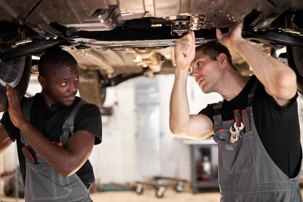 Dos interracial diversos mecánicos comprobar coche inferior — Foto de Stock