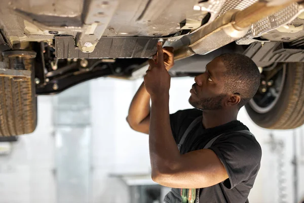 Joven mecánico de coche en coche de control uniforme en el servicio de automóvil con vehículo levantado — Foto de Stock