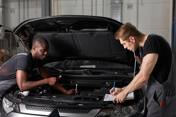 Dos diversos mecánicos automotrices que cooperan mientras que examinan funcionamiento del motor del coche en taller de reparación auto — Foto de Stock