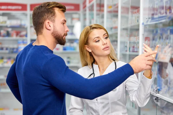 Junge Apothekerin hilft männlichen Kunden bei der Auswahl verschreibungspflichtiger Medikamente aus dem Regal — Stockfoto