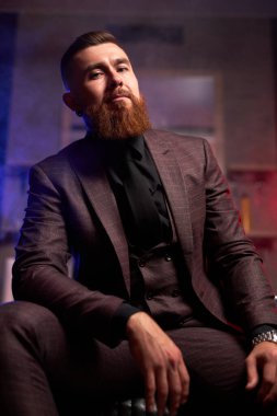 Smokinli yakışıklı genç sakallı adam karanlık odada yalnız oturuyor.