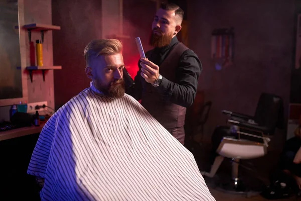 Acconciatore maschile sicuro tagliare i capelli all'uomo elegante elegante seduto nel salone — Foto Stock