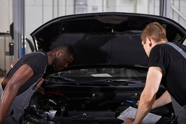 汽车修理厂汽车发动机性能检测中的两个不同的汽车机械师合作 — 图库照片