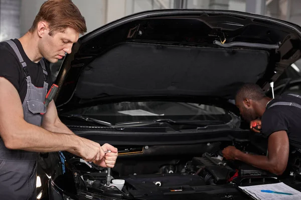 Dos diversos mecánicos automotrices que cooperan mientras que examinan funcionamiento del motor del coche en taller de reparación auto — Foto de Stock