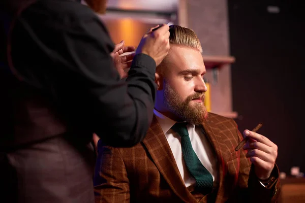 Стильний чоловік насолоджується стрижкою на перукарні, він курить під час процесу різання — стокове фото