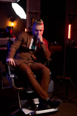 Smokinli yakışıklı genç sakallı adam karanlık odada yalnız oturuyor.