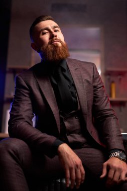 Genç, beyaz, yakışıklı, sakallı adam, karanlık dumanlı bir odada kanepede oturuyor.