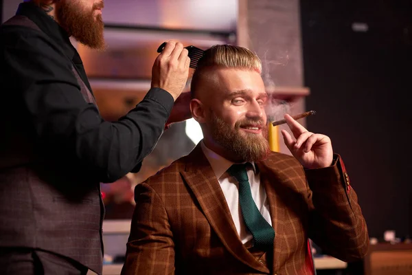Barbeiro cortar cabelo de seu cliente sentado em poltrona e fumar charuto — Fotografia de Stock
