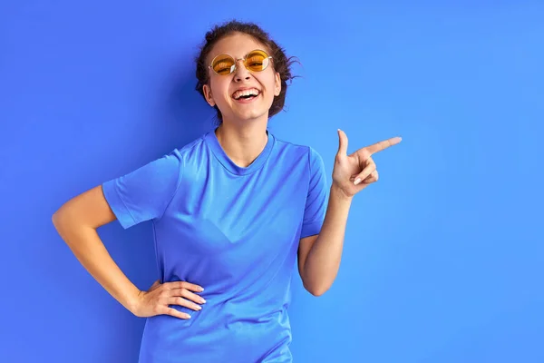 Alegre adolescente en camiseta posando en la cámara, señalando los dedos a un lado —  Fotos de Stock