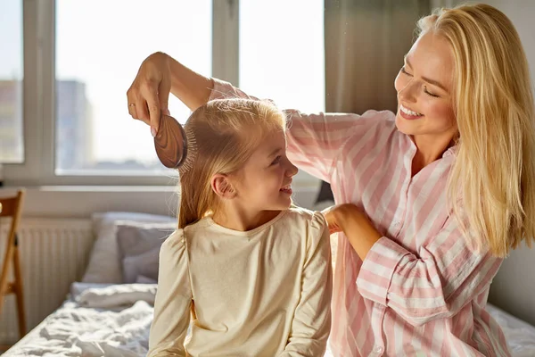 Feliz familia caucásica. Madre está peinando el pelo de sus hijas sentado en la cama en la habitación —  Fotos de Stock